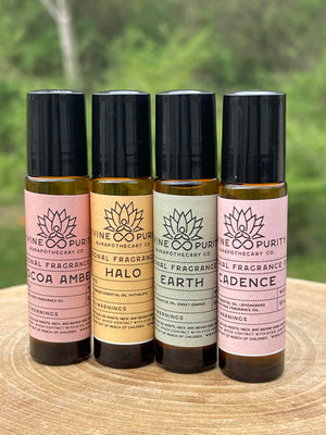 Four 10 mL amber roller ball bottles on wood with a green outdoor background. Divine Purity Personal Fragrance Oil in Cocoa Amber, Halo, Earth, and Cadence. Labels in dusty rose, gold, and sage gray.